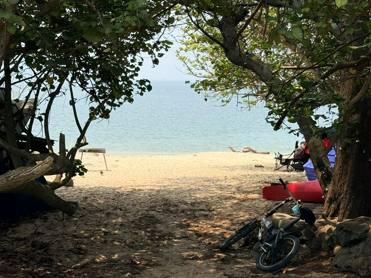 Lamma Island, Hong Kong — a gap in the tree shade opening onto the beach, a bicycle parked on the sand, pale blue sea beyond