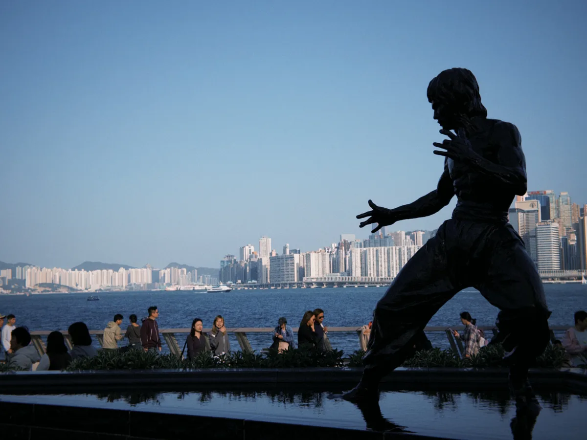 The Bruce Lee statue in silhouette by the water, dense high-rises on the opposite shore